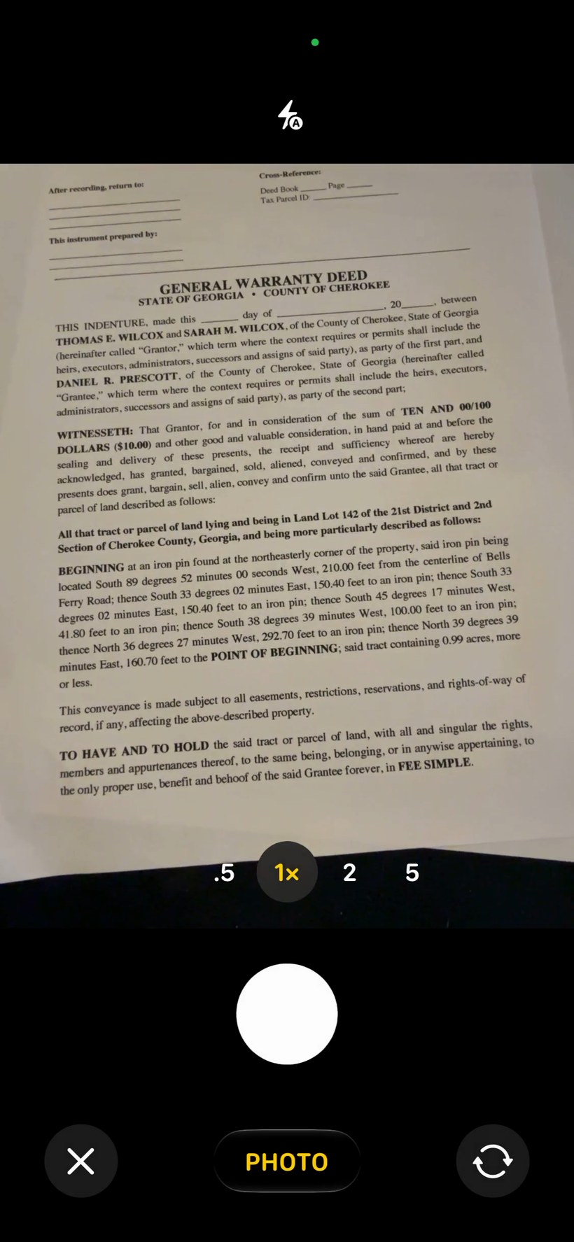 iPhone camera view aimed at a printed deed document — a sample deed is visible in the frame with the iOS camera shutter controls at the bottom